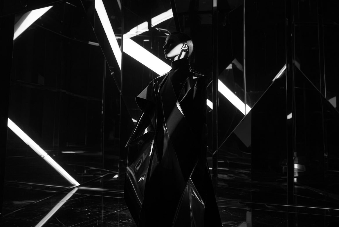 Cloaked figure walking a corridor of angled mirrors and white light, evoking exploratory creative work